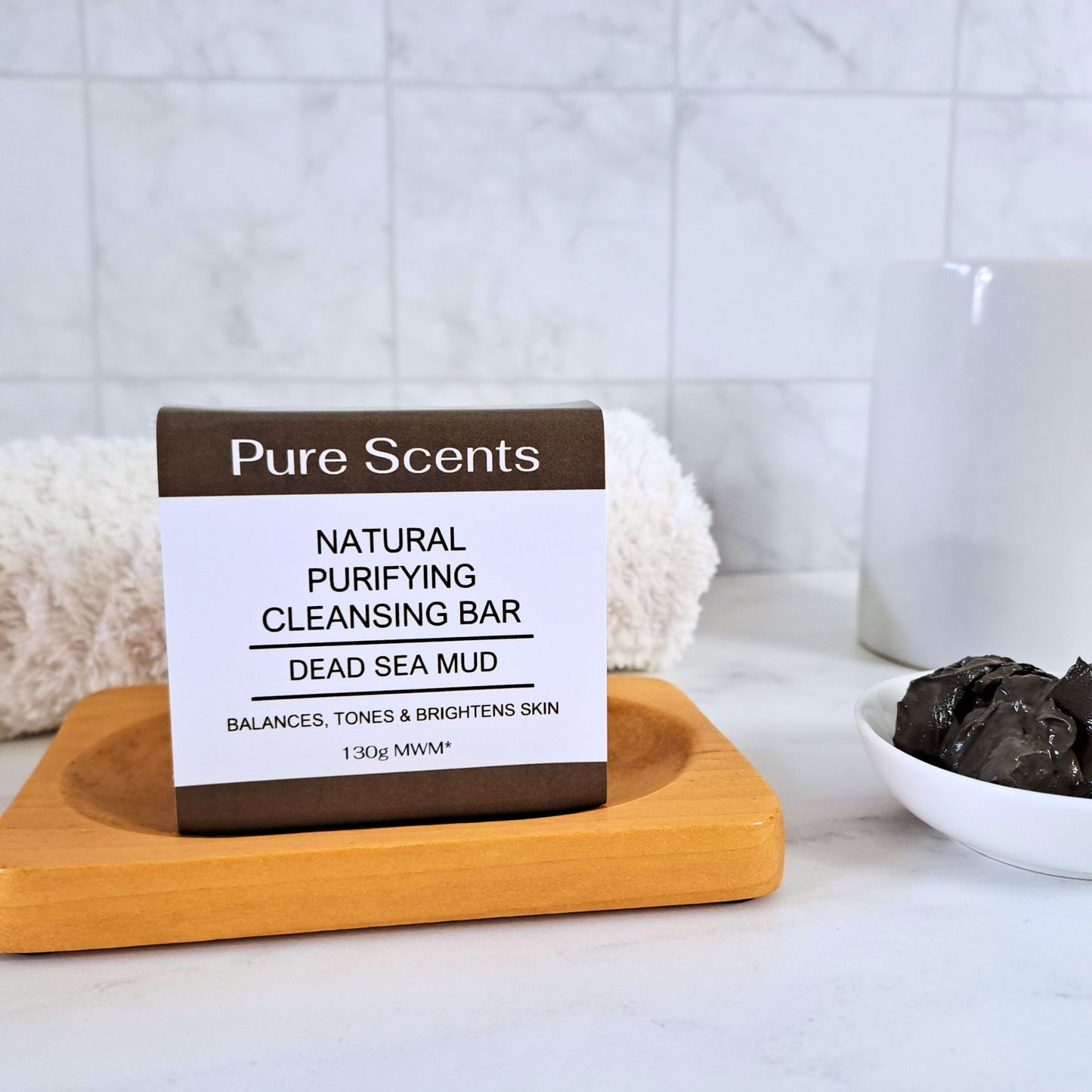 a bar of Dead Sea Mud soap sitting on top of a wooden soap dish surrounded by bathroom accessories with a marble tiled background.
