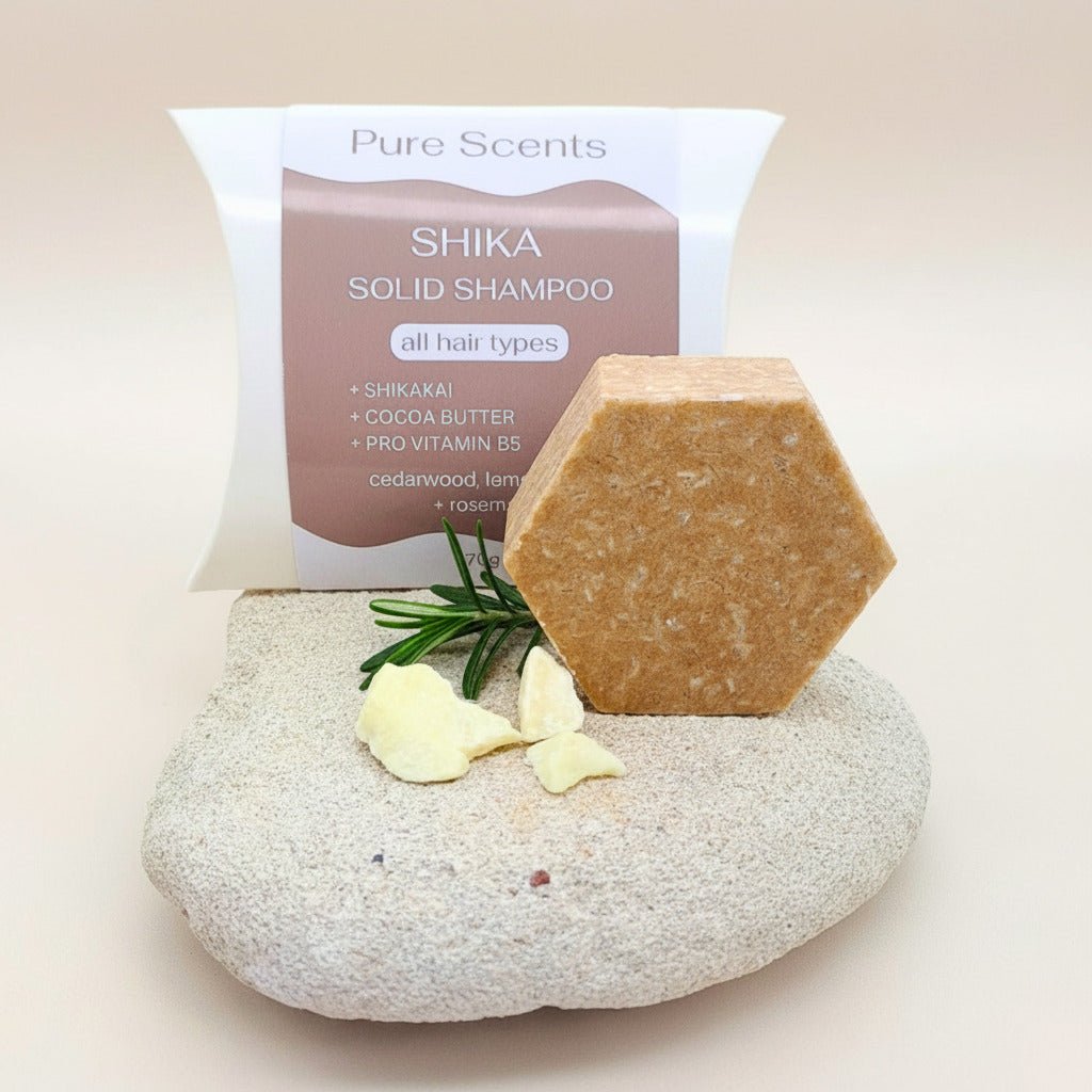 A hexagonal bar of Pure Scents Shika Solid Shampoo stands on a textured stone with a spig of rosemary and pieces of raw cocoa butter, with Pure Scents branded pillow box behind it on a light neutral background.